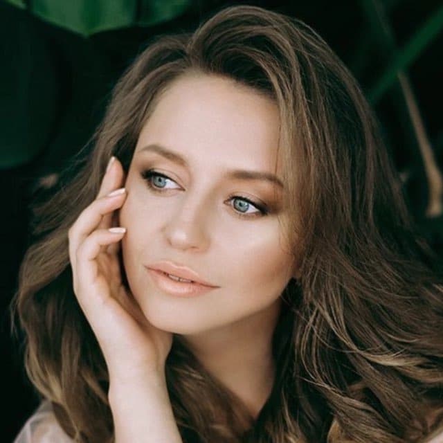 Woman with long wavy brown hair and blue eyes looking away with hand on cheek.