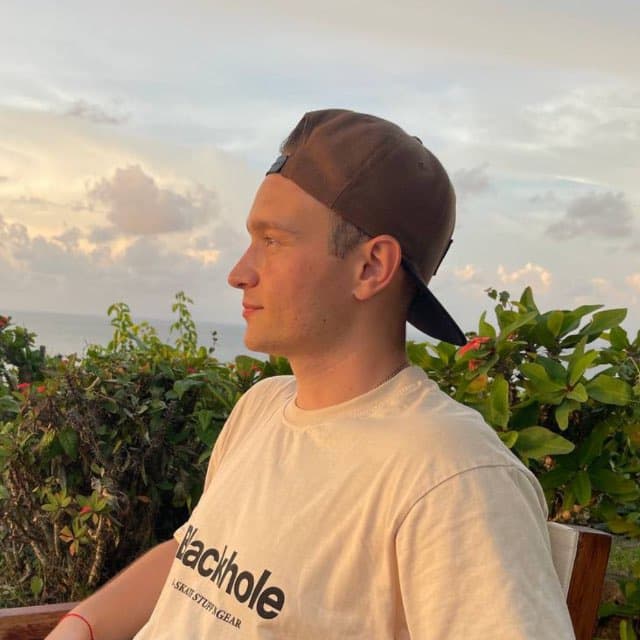 Man in a brown cap and cream shirt overlooking the ocean at sunset.